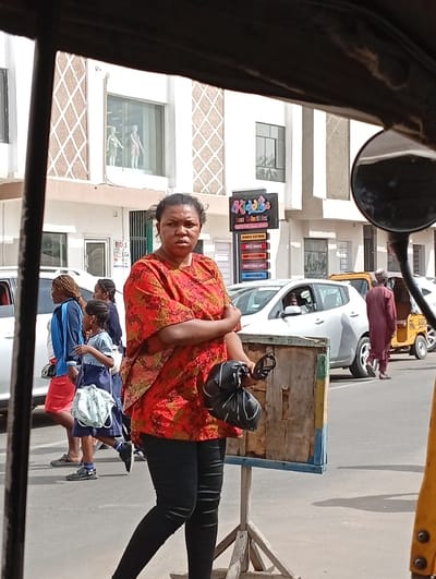 The image depicts an urban street scene in Sabon Gari East, Nigeria, captured from inside a vehicle. In the immediate foreground, dark framing from the capture vehicle is visible, with a circular side mirror on the right.

At the center, a woman with dark hair, wearing a red and orange patterned short-sleeved top and black leggings, stands facing the camera with a neutral expression. She holds a black, wrapped object in her left hand, which is held across her midsection, while her right arm is bent and resting on her left. To her right, a simple wooden stand, constructed from light-colored planks with blue accents on its lower edges, is positioned.

In the mid-ground, a white four-door sedan (identifiable as a Nissan Leaf) is parked or moving slowly. A person is visible in the driver's seat. Further right and behind the sedan, a man in a maroon traditional outfit walks from left to right. To the left of the central woman, two pedestrians are walking from left to right: a woman in a blue long-sleeved top and a red skirt, carrying a bag, followed by a young girl in a blue and white school uniform, carrying a light-colored bag.

The background features a multi-story white building with distinctive brown diamond-patterned panels on its upper facade. Two mannequins, one appearing male and one female, are visible in a large window on the second floor. On the ground floor, a black vertical sign for "Kiddies Lux Collectibles" lists categories such as 'Kids Clothing', 'Baby Essentials', 'Toys & Shoes', 'Party Pack & Gift', and 'Accessories'. Another yellow tricycle (Keke NAPEP) is partially visible on the far right of the frame. The scene is illuminated by bright daylight, casting defined shadows on the dry asphalt road.