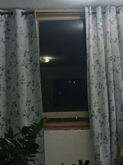 The image shows a window with floral patterned curtains on either side. The window is dark, suggesting it is night or the interior is not lit. There is a faint reflection of a light fixture on the ceiling visible in the window. A plant is partially visible in the lower left corner. The scene is indoors, likely a residential setting. No specific location in Valencia, Venezuela can be determined from the image.