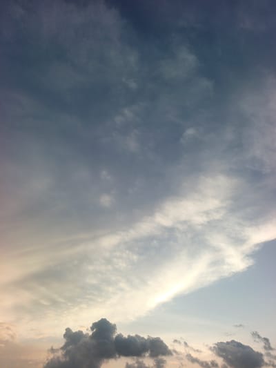 The image depicts an atmospheric sky scene over Rafin Bauna, Nigeria, likely captured during either dawn or dusk. The sky presents a distinct gradient, transitioning from a deep, dark blue-gray at the top to a lighter, pale blue-gray in the mid-section. Towards the bottom of the frame, a warm, soft yellow and orange glow is visible, indicating the presence of the sun near the horizon, just outside or at the very edge of the view.
Various cloud formations are present. The upper and middle portions of the sky feature diffuse, wispy white clouds, some appearing as elongated streaks and others as more scattered, lighter patches. These high-level clouds are less dense and allow some of the sky color to show through. Along the lower horizon, a band of darker, denser, and more textured clouds is visible. These clouds, potentially cumulus or stratocumulus, are silhouetted against the brighter lower sky, exhibiting irregular shapes and greater volume. There are no discernible landmasses, structures, people, animals, or man-made objects within the frame; the image focuses exclusively on the sky and its cloud cover.
