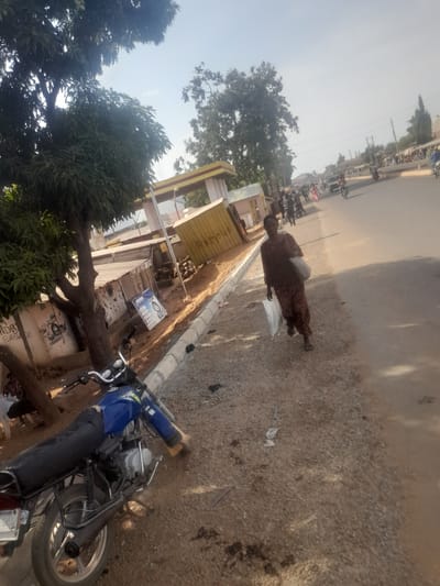 The image depicts a street scene in Ube, Nigeria, during daylight hours. In the foreground, a blue commuter motorcycle is parked on a dirt and gravel shoulder on the left, facing towards the right with its front wheel slightly turned. Its black seat is visible, and the rear tire and exhaust are distinguishable. To the right of the motorcycle, a person is walking towards the viewer, clad in dark, patterned clothing (possibly brown or maroon), carrying a white plastic bag in their right hand and a larger, light-colored bag over their left shoulder.
Behind the motorcycle and the walking individual, a partially completed concrete curb separates the gravel shoulder from a paved road. To the far left, a large tree with dense green foliage provides shade. Adjacent to the tree, behind the motorcycle, are several structures including a yellow container-like building with "REMON" partially visible on its side, and other buildings with corrugated metal roofs. Graffiti resembling a washing machine and "LAUNDRY SERVICE" is visible on one wall, while another sign depicting tire-related services is present further along the shoulder.
The paved road extends into the background, where multiple individuals are visible walking and operating motorcycles. A few vehicles, including what appears to be a dark-colored car or truck, are also on the road. The sky above is clear and bright, indicating a sunny day. The overall environment suggests an active, semi-urban or commercial area.