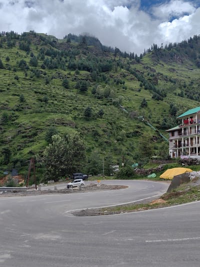 The image shows a winding asphalt road in Palchan, India, with a steep, green, tree-covered mountainside on the left. A multi-story building with a green roof, balconies, and laundry hanging out is visible on the right side of the road. A large yellow tarp is spread out near the building, and piles of firewood are stacked behind it. Two vehicles, a white car and a dark SUV, are parked on the side of the road near the base of the mountain. A power pole with wires stands on the left side of the road. The sky is cloudy with patches of blue.