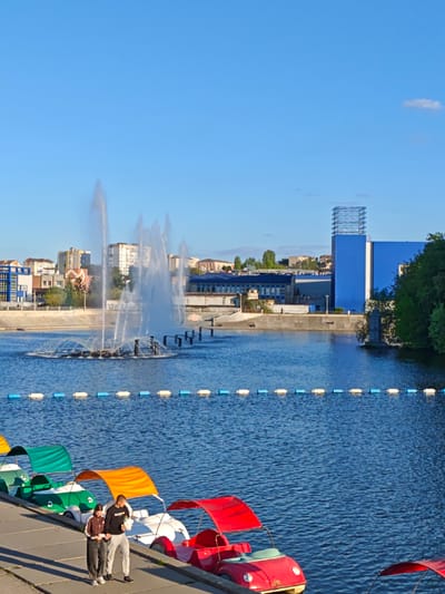 The image shows a waterfront scene in Vinnytsia, Ukraine. A body of water, likely the Southern Buh river, features a fountain in the middle. A line of colorful paddle boats, resembling small cars, are docked along the concrete bank. A couple, a man and a woman, are walking along the promenade next to the boats. The man wears a dark sweatshirt and light-colored pants. The woman is in a track suit. Buildings are visible in the background, including a tall blue structure with a metal framework on top. The sky is clear and blue.