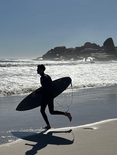Here is a detailed description of the image:
A silhouette of a surfer standing on a beach with his surfboard in Hout Bay, South Africa. The surfer is wearing a black wetsuit. Waves are approaching the beach and reflecting sunlight. Behind the surfer, to the left, are large rocks on the beach. The sky is a clear pale blue. The shadow of the surfer and the surfboard is visible on the wet sand.