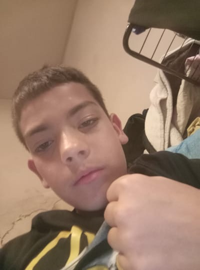 A close-up, upward-angled photograph shows a person with light brown skin, short, dark brown hair, and brown eyes. The person's face is centered in the frame, with their nose and mouth slightly prominent. They are wearing a black hoodie with yellow stripes on the shoulder and arm, which is visible on the left side of the image. The person's left hand is clenched into a fist and positioned near their chest.

Behind the person, slightly out of focus, is a messy environment. There are blankets or clothes stacked on top of each other, and a dark-colored metal frame, possibly part of a bed or furniture. The background wall is a neutral, beige color.

The location in Dolna Kremena, Bulgaria cannot be determined from this image alone. No specific elements in the image suggest this location.