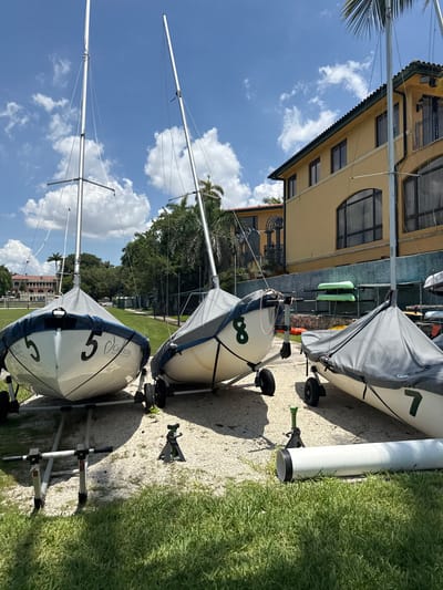 The image shows a sunny day in Miami, United States of America. The scene features several small sailboats on trailers, parked on a gravel surface next to a grassy area. Behind the sailboats, a building with a yellow facade and dark windows is visible. The sky is a bright blue with scattered white clouds, indicating good weather. Two sailboats are clearly visible in the foreground, each covered with a gray tarp. The front sailboat has the number "5" painted in black on its side. The second sailboat from the front has the number "8" painted on it. Several palm trees and lush green vegetation are present in the background, further suggesting a warm, tropical location. The image appears to be taken during daylight hours. The overall mood is serene, with the boats ready for use or being stored. There is a white cylindrical object, possibly a storage container or part of a dock structure, to the right of the second sailboat. Text is visible on the tarp of the second sailboat from the front, but it is difficult to read clearly. However, on the front sailboat, the words "CARILLON" and "MIAMI" are visible.