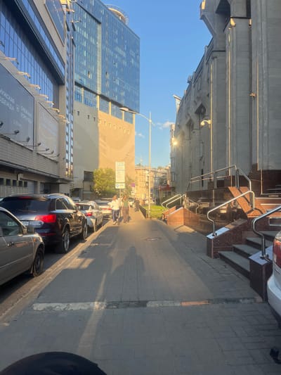 The image depicts an urban street scene in Voronezh, Russia. In the foreground, several cars are parked along the side of a paved sidewalk. The street is lined with modern buildings, including a prominent glass-fronted office building and a multi-story structure with storefronts. 

The scene appears to be captured during the late afternoon or early evening, as indicated by the long shadows cast across the sidewalk and the warm sunlight illuminating the buildings. The sky is clear and blue, suggesting fair weather. 

Visible text includes signage for "OMTA", "СНЕЖНАЯ КОРОЛЕВА" (Snow Queen), "2 ЭТАЖ" (2nd Floor), and "Леонардо хобби-гипермаркет" (Leonardo Hobby Hypermarket). License plates are also partially visible. 

A few people are walking on the sidewalk in the middle ground, suggesting a normal day in the city. The overall impression is one of a bustling urban environment bathed in the golden light of the setting sun.
