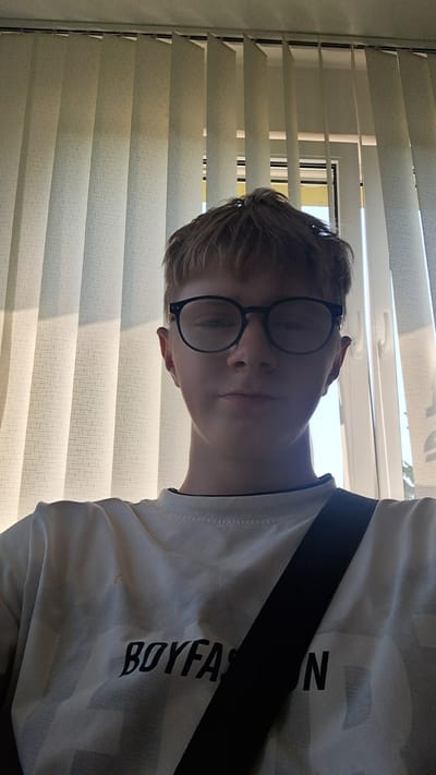 This media file is a selfie of a young person wearing glasses and a white t-shirt with a black strap across it. The t-shirt has black text on it. The person is looking down and slightly to the side. The background consists of blinds, suggesting an indoor setting, possibly a room. The lighting indicates it is daytime, likely sunny due to the brightness through the blinds. The overall mood appears casual and relaxed. The text visible on the t-shirt reads "BOYFAST".