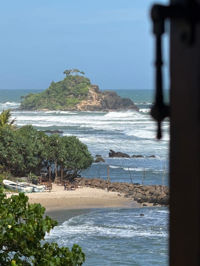 The image captures a coastal scene in Goviyapana, Sri Lanka. In the distance, a small, rocky island covered in lush green vegetation and topped with a few trees rises from the sea. Waves crash against its shores. Closer to the foreground, a sandy beach is visible, lined with several tables and chairs, suggesting an outdoor dining area. Two small boats are pulled up onto the sand near the tables. Dense green foliage, including palm trees and other leafy plants, frames the left side of the image and extends towards the beach. The ocean is a deep blue, with white waves breaking and receding. The sky above is clear and blue. A dark, out-of-focus frame is present on the right side of the image, partially obscuring the view. No people are visible in the scene.