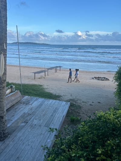 This media file captures a serene beach scene in Peleana, Sri Lanka, likely during the late afternoon or early evening. The image is oriented vertically.
The foreground features a rustic wooden deck, with weathered planks laid out at an angle. To the left, a rough-textured, dark surface, possibly a tree trunk or a stone structure, is visible, with some rope wrapped around it. Lush green foliage hangs from the upper right corner, casting shadows on the sandy beach.
The middle ground reveals a sandy beach with gentle waves lapping the shore. Two men are walking side-by-side on the sand. One is wearing a white t-shirt and khaki shorts, while the other wears a blue t-shirt and khaki shorts. They appear to be casually strolling. Further down the beach, several empty wooden lounge chairs with slatted tops are arranged in a row, suggesting a resting area for beachgoers. A peculiar arrangement of rocks, forming a linear pattern, is visible in the sand.
The background showcases the ocean, with distinct lines of waves rolling towards the shore. The water is a serene blue, reflecting the sky. In the far distance, a faint outline of land and structures can be seen along the horizon. The sky is a vibrant blue, with scattered clouds, some fluffy and white, others darker and more dramatic, hinting at possible atmospheric conditions. The overall mood is peaceful and tranquil, evoking a sense of calm vacation or relaxation. There is no visible text in the image.