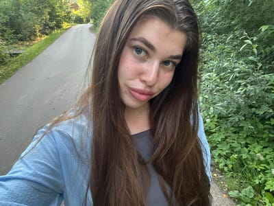A young woman with long, dark brown hair poses for a selfie in an outdoor setting. She is wearing a light blue textured top over a dark grey t-shirt. Her face is the primary focus, with her head tilted slightly. She has blue-grey eyes, light eyebrows, and pink lips. Her skin appears fair with some minor blemishes.

The background includes lush green foliage to her left and a paved pathway to her right, suggesting a park or rural road. The lighting indicates it is daytime, likely with bright sunlight casting some shadows, especially on the pathway. The overall mood seems casual and relaxed. There is no visible text in the image. The location is identified as Shevlyagino, Russia.