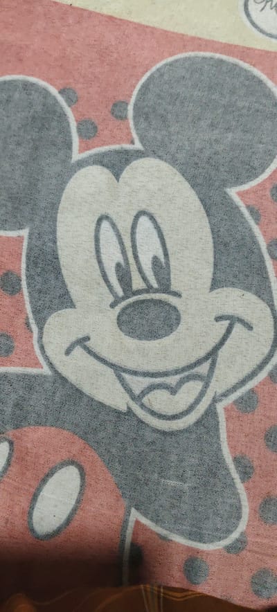 This is a close-up shot of a patterned fabric, likely a bedsheet or blanket, captured in Paloh Teungoh, Indonesia. The main subject is a large, smiling Mickey Mouse head, depicted in light gray with dark gray outlines for his face, prominent ears, eyes, and nose. His expression is cheerful, featuring a wide smile that reveals his tongue, and his eyes are wide open.
Surrounding Mickey and forming part of the overall pattern are areas of pink fabric adorned with dark gray polka dots. Elements of other character designs or repetitions of the pattern, such as a pink polka-dotted section with a white outline in the bottom left, are also partially visible. The fabric exhibits a clear woven texture, and the colors appear slightly muted or faded, suggesting age or a particular print style. No specific activity or event is taking place, as the image is a static depiction of the textile. There is no visible text. The time of day and weather conditions are not discernible from the photograph.