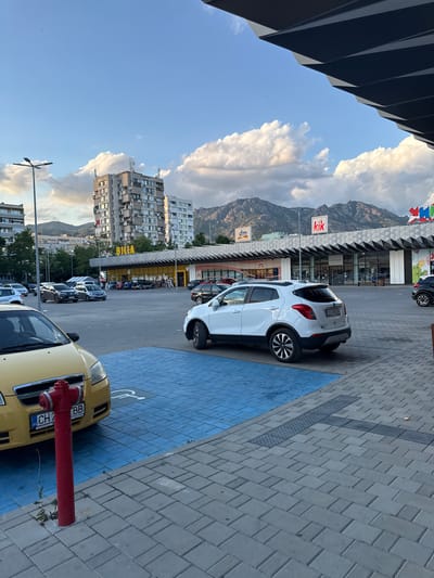 The image shows a parking lot in front of a commercial building with multiple storefronts, including "BILLA", "dm", and "kik". Mountains are visible in the background under a cloudy sky. In the foreground, a yellow car is parked, and a red fire hydrant stands next to it. A white SUV is parked in a designated blue parking spot with a disabled symbol. The scene appears to be in the city of Sliven, Bulgaria, given the context of the available storefronts. The time of day appears to be late afternoon or early evening, as indicated by the lighting and the sky.