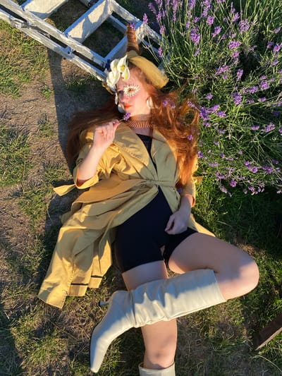 A woman with long, auburn hair lies on her back in a grassy field dotted with lavender. She is wearing a yellow trench coat, black shorts, and cream-colored knee-high boots. Her head rests on a silver ladder, and a wide-brimmed hat covers her forehead. White calla lilies and a daisy are arranged on her face, with the daisy placed over her eye. She holds a small purple flower in her right hand, bringing it towards her face. The sunlight suggests it is late afternoon, casting long shadows. The scene evokes a sense of tranquility and connection with nature. No visible text is present in the image.