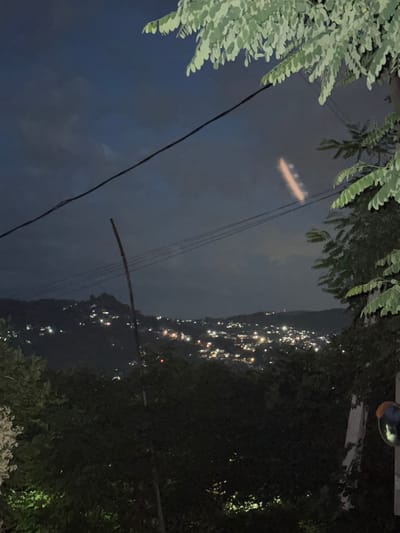 The image captures a night scene overlooking a town with scattered lights. In the foreground, dark foliage from trees dominates the lower and right portions of the frame, with a visible telephone pole on the far right. Overhead, power lines stretch across the dark sky, with a faint, glowing streak of light visible in the upper center, possibly from an aircraft or atmospheric phenomenon. In the mid-ground, the lights of a town are spread across a hillside, suggesting a settlement in a hilly or mountainous terrain. The sky is dark and cloudy. The location is identified as Baranovka, Russia.