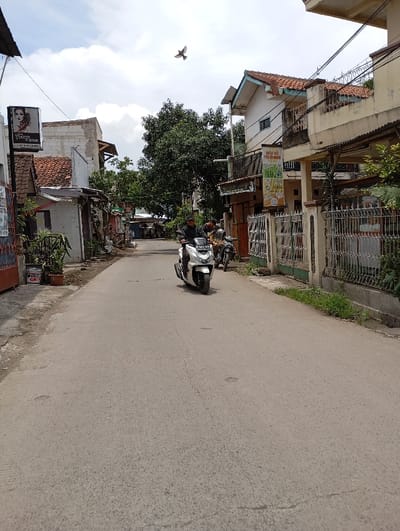 The scene is a street in Katapang, Indonesia, located at -6.985 latitude and 107.557 longitude. It's a residential street with buildings on both sides. There is a man riding a white scooter in the middle of the street. The scooter is moving towards the right side of the image. There is a second scooter parked on the right side of the image with a man sitting on it. The man on the scooter is wearing a helmet and a white shirt. The road is paved and empty, except for the scooter. The sky is clear with some clouds. A bird is flying in the sky. The surrounding buildings are residential, and some have metal fences. There is a sign on the left side of the image that says "Hair Salon". There is a sign on the right side of the image that says "DATO AC PALOMA". There is a tree on the left side of the image, and another tree on the right side. There are some plants growing on the side of the road. There is a white building on the right side of the image with a large metal fence. There is a small group of buildings in the left side of the image, appearing to be a commercial area. The street appears to be relatively quiet.