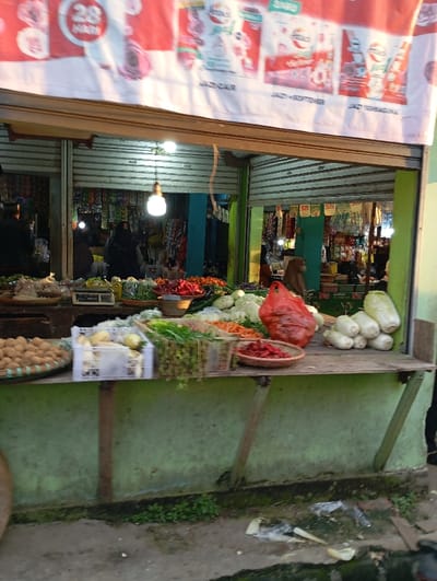 The image shows a street-level view of an outdoor market stall in Kutawaringin, Indonesia, located at the coordinates -6.953 latitude and 107.524 longitude. The stall is positioned in front of a larger building with a corrugated metal roof. Various products are displayed on the wooden counter: a basket of round brown fruits, a plastic crate of white vegetables, a basket of green vegetables, a basket of carrots, and a basket of red chili peppers. A red plastic bag containing unknown contents sits on the counter, adjacent to a stack of white vegetables. Several people stand behind the counter, but their faces are not visible. The stall is partially covered by a red and white awning with the words "JAGOR" and "JACK SOFTENER" printed on it. The ground beneath the stall is a mixture of dirt and overgrown grass. A few plastic bags and pieces of debris are scattered on the ground in the foreground.