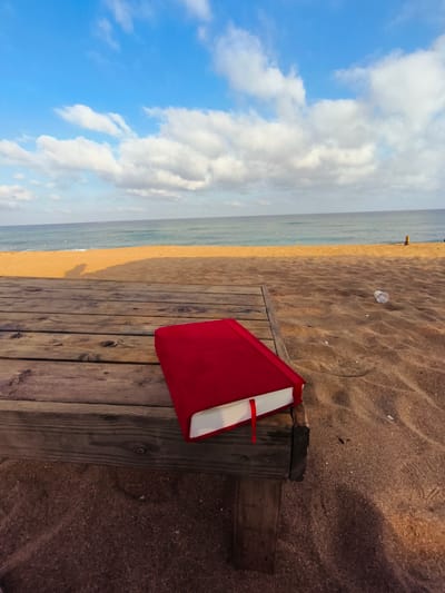 A red-covered book rests on a wooden platform on the sandy beach of Nahariyya, Israel, with the Mediterranean Sea and a blue sky with scattered white clouds in the background. The platform is constructed of weathered wooden planks, and the sand is fine-grained with visible footprints and some debris, including a discarded plastic cup to the right of the book. The book is closed, with a red ribbon bookmark hanging from the side, and the edges of its white pages are visible. The sea is calm, with a gentle wave line meeting the sandy shore. The lighting suggests it is late afternoon or early morning, with a shadow of what appears to be a person cast on the sand to the left of the platform.