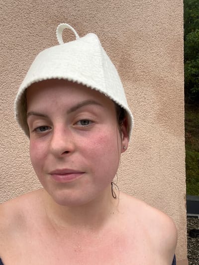 A close-up, portrait-oriented photograph depicts a single adult female in Staudernheim, Germany. The subject is centrally positioned, facing the camera directly with a neutral expression and light-colored irises. She wears a white, conical felt sauna hat featuring a scalloped brim and a small loop at its apex. Her skin, particularly across the face, neck, and visible upper chest and shoulders, appears flushed and reddened, covered with numerous small water droplets. Dark, damp hair is visible beneath the hat, with strands adhering to the right side of her face (viewer's left). A small, clear stud earring is present in her right earlobe (viewer's left). She appears bare-shouldered or minimally clothed, with a dark garment strap visible on her left side (viewer's right). The background consists primarily of a textured, light beige stucco wall. A narrow vertical section on the far right reveals green foliage, indicating an outdoor or garden setting adjacent to a structure. The scene is illuminated by diffused natural light.