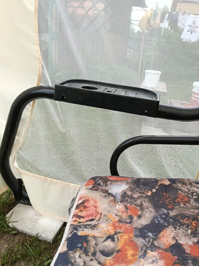 The image shows a close-up view of a swing in what appears to be a backyard setting, likely in Красный Маяк, Russia. The swing has a black metal frame with a tray attachment on the armrest. The seat cushion is floral patterned with shades of orange, brown, and gray. A light beige fabric panel hangs from the frame. A fine mesh netting surrounds the swing. In the background, clothes are hanging on a clothesline. A white bucket sits on a surface. The ground around the swing has grass and concrete tiles.