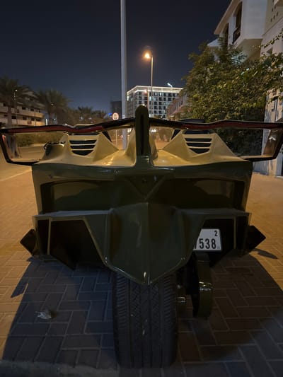 Centered is a military-green vehicle with a spare tire mounted on the rear. A license plate displaying "5382" is visible. The vehicle is parked on a brick-paved surface, likely a sidewalk or street. The background features buildings, including one that appears to be residential, with palm trees in the distance. A street light illuminates the nighttime scene.