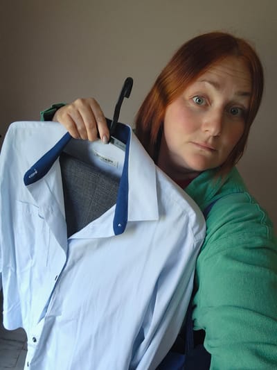 A woman with red hair and green eyes is holding a light blue dress shirt on a black plastic hanger. The shirt has a dark blue trim on the collar and a small dark patch visible inside the collar. She is wearing a green long-sleeved shirt. The background is a plain light brown wall. The image does not contain enough information to determine the location of Markovskii, Russia.