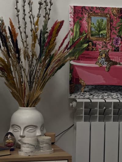 The image depicts an interior scene. On a white wall hangs a decorative panel featuring a floral and leopard print design with a framed landscape painting within the larger design. To the right of the panel, a white radiator is mounted on the wall. Below the panel, a white vase shaped like a human head sits on a wooden surface. The vase contains dried wheat stalks and pussy willow branches. Adjacent to the vase, there are two marble-patterned cups and a glass decorative object. The location is not discernible as Zovuni, Armenia, based solely on the provided image. The image provides no information about people, actions, or interactions.