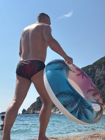This media file captures a person holding an inflatable ring at Mirište, Montenegro. The subject, a man, is wearing patterned swimming briefs and appears to be on a beach. He is seen from behind, with his back and buttocks prominently featured. The inflatable ring has a tropical design with palm trees and a gradient of colors ranging from purple to blue.
The scene is set against a clear blue sky, suggesting a sunny day. To the right of the subject, the turquoise waters of the sea are visible, with gentle waves and sunlight reflecting off the surface. The shoreline, composed of small pebbles and sand, is also visible, with some debris scattered along it. In the background, a rocky cliff face adds to the natural beauty of the location. The lighting indicates it is daytime, likely late morning or early afternoon, given the brightness of the sun.
The person's pose, with one arm extended and holding the inflatable ring, suggests an action or anticipation of an activity, possibly entering the water or preparing to play. The overall mood appears relaxed and recreational, fitting for a beach environment. No text is visible in the media file.
