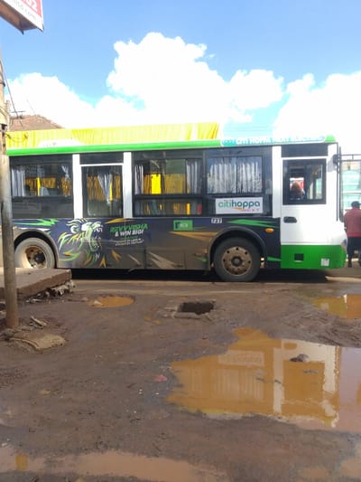A white and green bus with the words "Citi Hoppa Next Electric" on the side is parked on the side of a dirt road in Athi River, Kenya. The bus is facing to the right of the image and has a number "727" visible on the side. There is a puddle of brown water in the foreground, reflecting the bus and the sky. The bus has a large graphic of a lion with the words "Revvysha & Win Big" on the side. There are two people visible in the bus's windows. The bus is parked in front of a building with a yellow awning. The sky is blue with white clouds. The road is muddy and there are several potholes.
