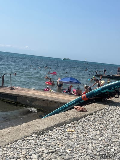 The image depicts a busy beach scene in Blagoveshchenskaya, Russia, on a sunny day. The sea is calm with a clear blue sky. Many people are swimming and relaxing in the water. Some individuals are using inflatable rings for leisure. On the shore, a concrete pier runs along the water's edge. A blue umbrella provides shade for a group of people sitting on the pier. In the foreground, a stand-up paddleboard is resting on the pebble-strewn beach. In the distance, a boat is visible on the sea, suggesting recreational boating activity. The overall atmosphere is one of a lively summer day at the beach.