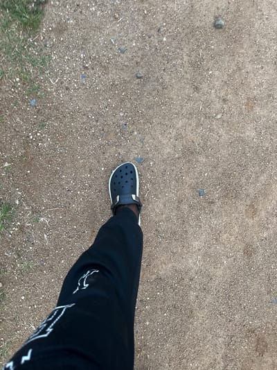 The media file shows a person's leg and foot wearing black Crocs and black pants. The Crocs are dark and appear to have a white trim. The pants are also black and have white graphic designs on them, possibly a logo. The person is standing on a ground covered with small stones and dirt. Some sparse dry grass is visible in the lower part of the frame. The scene appears to be outdoors, given the ground texture and the natural lighting. The time of day is likely daytime, as indicated by the brightness of the scene. The weather seems to be dry, with no signs of rain or wetness. The image provides no clear indication of the specific location within Gwong, Nigeria, beyond it being an outdoor area with a rough, unpaved surface. The activity depicted is simply the person standing or walking, as suggested by the angled perspective of the leg and foot. There is no explicit emotional expression conveyed due to the focus on the lower body and the ground. Some text is visible on the Crocs, appearing as a white logo on the side strap, and also on the pants as white graphic patterns.