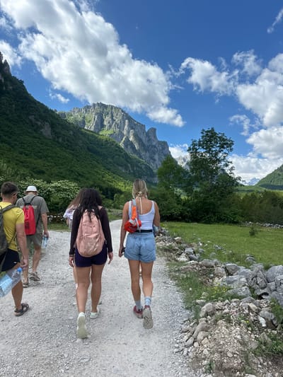 A group of five individuals is captured from behind, walking along a gravel path in a mountainous, rural landscape. The group consists of four adults and one person whose age is indeterminate but appears to be an adult. Two women in the foreground are wearing backpacks. The woman on the right wears denim shorts, a white tank top, and a red pack. The woman next to her wears dark shorts and a pink backpack. Behind them, a man in a light-colored hat and a green t-shirt with a red backpack walks with a water bottle. To the left of him, another man in a yellow t-shirt and dark shorts carries a water bottle. The path is flanked by green grass and rocky terrain. Towering, verdant mountains rise in the background under a bright blue sky with scattered white clouds. The image is taken in the vicinity of Gusinje, Montenegro, a location known for its dramatic mountain scenery and natural beauty.