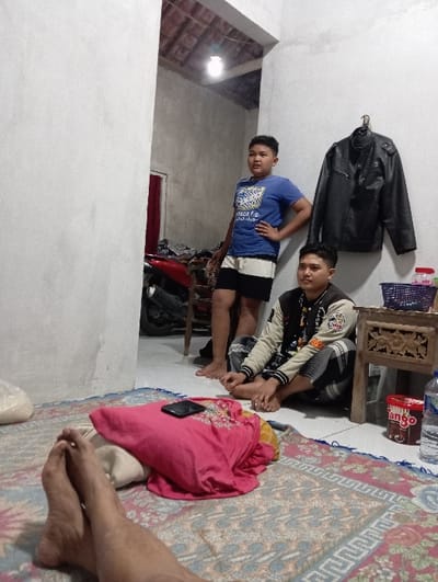 The image depicts an indoor scene in Bulukandang, Indonesia, featuring three individuals within a modestly furnished room. The floor is covered by a patterned rug in shades of brown, blue, red, and beige. The walls are light-colored and unadorned. A single bare light bulb hangs from the ceiling. In the background, an open doorway reveals a darker area with a red motorcycle or scooter visible, and an exposed tiled roof structure overhead.
Two males are prominently featured. A standing male, appearing to be a juvenile, is positioned on the left side of the frame, facing slightly right with his left hand on his hip. He wears a blue t-shirt with a white graphic and dark shorts with a white stripe; he is barefoot. A seated male, appearing to be a young adult, is on the right, cross-legged on the rug, looking forward. He wears a cream-colored varsity-style jacket with dark sleeves and patterned grey pants; he is also barefoot. A black leather jacket hangs on a hook on the wall to the right of the standing male.
Adjacent to the seated male is a small, ornate carved wooden table holding a blue woven basket containing various small containers and a red-and-white container. On the floor nearby are a plastic bottle of water and a red-and-white cylindrical container labeled "Tango." In the immediate foreground, the bare legs and feet of a third individual are visible, suggesting they are reclining on the floor, likely operating the camera. A dark-colored smartphone rests on a folded piece of pink fabric or clothing on the rug near the foreground legs. A white plastic bag is partially visible on the far left of the foreground.