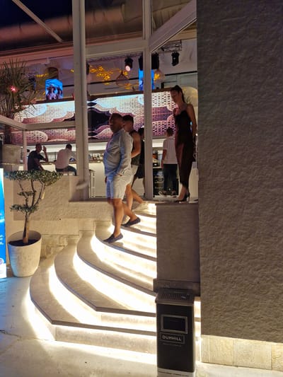 The image depicts an outdoor evening scene at a venue in Sveti Vlas, Bulgaria. In the foreground, a set of illuminated, curved stairs leads upwards. To the left of the stairs, a potted olive tree stands beside a concrete floor. Several people are visible. On the stairs, two men walk upwards; one wears a striped short-sleeved shirt and white shorts, and the other wears a dark t-shirt and shorts. A woman in a dark, flowing dress and high heels stands on a landing above and to the right of the stairs, facing slightly away from the camera. In the background, a bar area is visible with patrons and staff. Further back, large screens display images. A dark metal trash receptacle with a "DUNHILL" logo is positioned to the right of the stairs, near the bottom of the image. The overall environment suggests a lively outdoor establishment, possibly a restaurant or club, with ambient lighting contributing to the atmosphere.