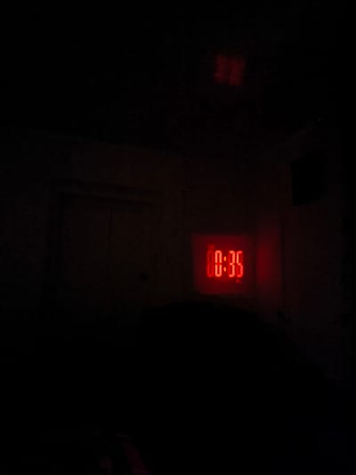 The image captures a dimly lit room, likely a bedroom, with a prominent red digital display showing the time "00:35". The display is part of an alarm clock, illuminated against the darkness. A dark, indistinct shape, possibly a person or bed, occupies the foreground. In the background, faint outlines of walls and possibly a door or window can be discerned, rendered in dark tones due to the low light conditions. The overall impression is one of a quiet, nocturnal scene. The location is specified as Kyzyl-Suu, Kyrgyzstan, suggesting an interior domestic setting within this city.