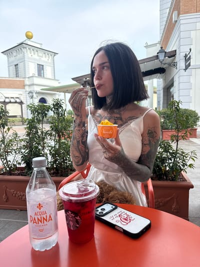 The image shows a woman with tattoos eating from a Venchi chocolate box at an outdoor table in Noventa di Piave, Italy. On the table is a bottle of Acqua Panna water, a Starbucks drink, and a phone in a "Not my circus, not my monkey" case. The background shows buildings and greenery typical of the Noventa di Piave area.