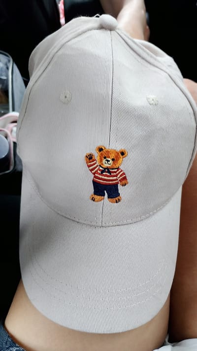 A close-up, top-down shot shows a light beige baseball cap worn by an unidentified person. The cap is embroidered with a small, orange-brown teddy bear wearing a striped shirt and dark blue pants. The teddy bear is waving with its right hand. The cap is made of ribbed fabric and has a stitched button on top. The background is out of focus and dark, suggesting an indoor setting, possibly a car interior given the presence of what appears to be a dark fabric seat behind the cap. The person's shoulder and upper arm are visible on the right side of the frame, showing light-toned skin. The image is tightly cropped, focusing primarily on the cap and the embroidery. There is no information in the image to associate it with Karlovo, Bulgaria.