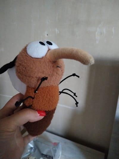 A plush toy mosquito is held in a person's hand against a neutral, lightly textured wall. The toy is predominantly brown and tan, with large white eyes and black pupils. It has a long, tan proboscis, black thread-like legs with small knots, and black and white wing-like appendages. The person's hand is visible at the bottom left of the frame, with a bright red painted fingernail. The background wall appears to be painted or stuccoed and has minor imperfections. The lighting is soft and appears to be from an indoor source. The scene does not contain any specific elements that can be used to determine its location in the city of Zovuni, Armenia.