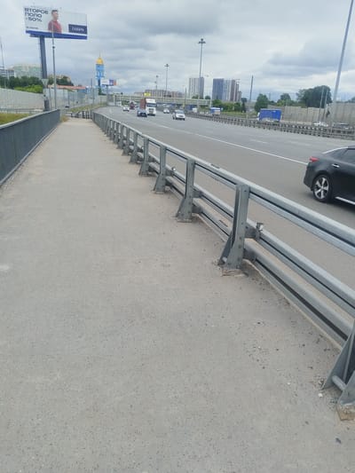 This image depicts a pedestrian walkway running alongside a highway in Mytishchi, Russia. A metal railing separates the walkway from the highway. Several vehicles, including cars and trucks, are visible on the highway. In the background, a blue and gold domed building, possibly a church, is prominent, along with apartment buildings and construction cranes. A billboard with a man's image and Russian text is also visible. The sky is overcast with clouds.