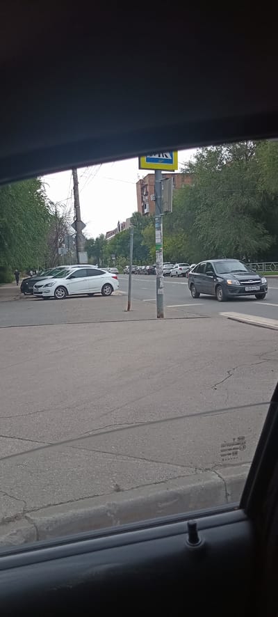 The image shows a street scene in Samara, Russia, viewed from inside a vehicle. The primary subjects are several parked cars on the side of a paved road. A white car is prominently displayed in the foreground, parked parallel to the curb. Behind it, a dark-colored van or SUV is also visible, along with other vehicles further down the street, suggesting a typical urban environment.
The scene is set on a clear day, with sunlight casting shadows and illuminating the road and vehicles. Lush green trees line the side of the road, indicating a time of year with foliage, likely spring or summer. In the background, buildings are visible, some appearing to be apartment complexes, consistent with a residential or mixed-use urban area.
A prominent pedestrian crossing sign with a yellow background and a blue pictogram of a person walking is positioned on a pole near the parked cars. This sign, along with the presence of multiple vehicles, suggests this is a public street and potentially an area with regular traffic. There are also power lines and street poles visible, adding to the urban context.
Notable details include the license plate on the dark-colored vehicle, which appears to read "X369TC 163". The overall impression is one of a calm, ordinary day in a Russian city, with no particular activity or event being depicted other than the general presence of parked cars on a street. The image is taken from a slightly elevated perspective, as if from the driver or passenger seat of a car.