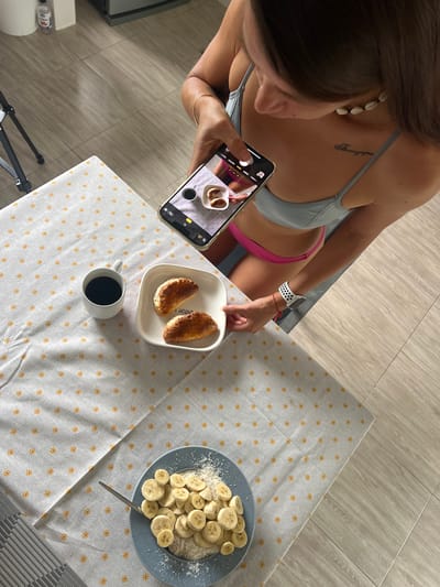 A woman is taking a photograph of breakfast with her smartphone. The breakfast consists of two croissants and a cup of coffee on a white plate with a patterned tablecloth. In the foreground, there is another plate with sliced bananas and shredded coconut, with a spoon resting on it. The woman is wearing a blue bikini top and pink bottoms. She has a seashell necklace and a watch on her left wrist. The scene appears to be in a kitchen or dining area with tiled flooring. The location is not visible in the image.