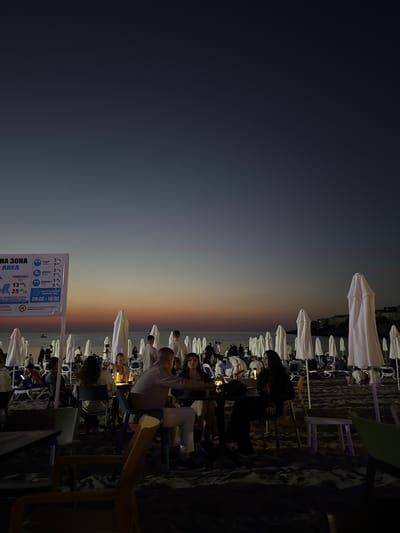 The image depicts a beach scene at dusk in Sozopol, Bulgaria. In the foreground, several tables and chairs are set up on the sandy beach, with people seated at some of them. The sky exhibits a gradient of colors from deep blue at the top to orange and pink near the horizon. Numerous white beach umbrellas are neatly arranged in rows behind the seating area. In the background, more people are visible, and a body of water can be seen with a faint coastline and buildings in the distance. A sign with text in Bulgarian and English is positioned on the left side of the frame, providing information about beach services. The overall atmosphere is tranquil, with the fading light creating a serene ambiance.