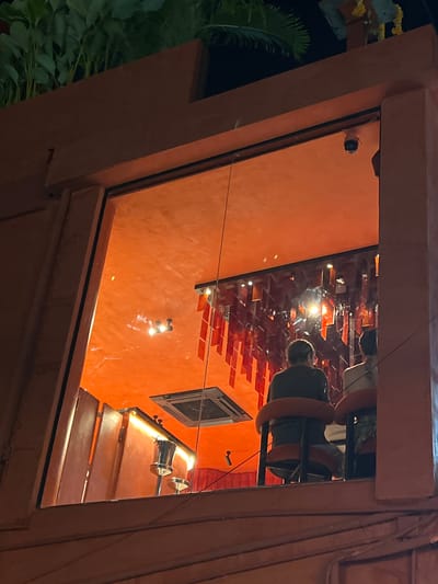 The image depicts an interior scene, viewed through a glass window or opening, with an orange-toned, textured wall as the dominant backdrop. Reflections on the glass reveal a ceiling adorned with numerous rectangular, orange tiles arranged in a visually interesting pattern. Within the reflections, parts of what appear to be people are visible, including a head and shoulders. One section of the reflection shows a dark object with orange bands, possibly part of a seating arrangement or light fixture. A small, dark, square-framed object, possibly a window or vent, is set into the orange wall. Below this, there's a shelf or ledge with wooden slats, and on this ledge are two metallic objects with a conical shape, reflecting light. The overall lighting suggests it is nighttime or a dimly lit interior space. The context provided places this scene in Baan Bo Phut, Thailand.