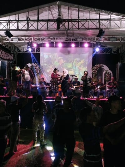 The image shows a stage performance taking place under a large tent.  Two women are singing on stage, one wearing a red shirt and the other wearing a light blue hijab. Behind them, a group of musicians are playing instruments. In the foreground, a group of people are watching the performance. The stage is lit with pink and white lights, and the tent is also lit with some white lights.  The location is near the city of Kecamatan Sambirejo, Indonesia,  at coordinates (latitude: -7.5, longitude: 111.087).  The environment is dark, with a grassy area surrounding the stage. The people in the image are all dressed casually and appear to be enjoying the performance.