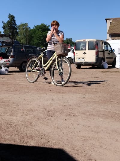 A woman with short brown hair, wearing a striped t-shirt and dark pants, is standing next to a light yellow bicycle on a dirt ground. She is holding a purple mobile phone to her right ear and appears to be talking on it. The bicycle has a wicker basket on the handlebars and a rack over the rear wheel. In the background, a dark grey car with its trunk open is parked on the left, and a beige van with its rear doors open is parked on the right. The environment is an outdoor area with a clear blue sky overhead. The scene appears to be in Irshansk, Ukraine, given the vehicle license plate which appears to be Ukrainian.
