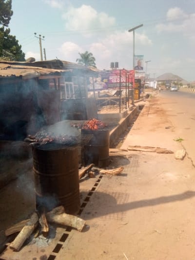 The scene depicts an outdoor food preparation area alongside a paved road in Agbani, Nigeria. In the foreground, two large, dark metal drums, possibly repurposed oil drums, are actively used as grills. The drum on the left emits significant white smoke, with charred wood visible at its base, indicating a fire. Both drums are topped with metal grates holding what appears to be grilled meat, some reddish-brown in color. Logs of wood are scattered around the base of the left drum and on the concrete pavement.
Adjacent to the grills, to the left, is a makeshift structure with a corrugated metal roof and walls, suggesting a street vendor stall. Behind the grilling area, a concrete drainage ditch runs parallel to the road. The immediate ground is a mix of concrete pavement and dirt.
Further down the road, to the right, a wide, paved road is visible, with one white vehicle driving away from the viewer and a solitary human figure walking in the distance. Utility poles with power lines are present on the left, and multiple tall, solar-powered streetlights line the road. Several billboards are erected along the roadside; one prominent blue billboard features a man's portrait with text "TUFU FOR GOVERNOR," and another large banner advertises "SPIRIT GLOBAL" and "SMOKED CHICKEN, FISH, & CATFISH." The sky above is light blue with scattered white clouds, indicating daylight.