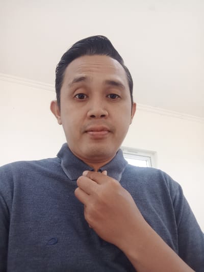 An Asian man wearing a dark blue polo shirt stands in front of a window, adjusting his collar with his right hand. He has short, dark hair styled upward and a slight smirk. The background is a bright, white-painted wall with a visible corner where it meets the ceiling. A window is slightly visible. The overall lighting is bright and even.
