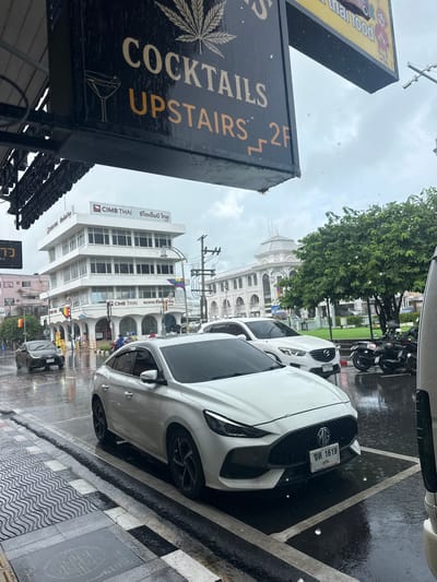 This media file depicts a street scene in Phuket City Municipality, Thailand, on a rainy day. The foreground features a dark sign with text and an illustration of a martini glass, partially obscured by water droplets. The text on the sign reads "COCKTAILS UPSTAIRS 2F".

Further into the scene, several vehicles are visible, including a white MG car with a license plate that reads "ขต 1619 ภูเก็ต PHUKETPIYA MG". The street is wet and reflective, indicating recent rainfall, with visible water on the pavement and the vehicles. In the background, a white building with arched windows and a domed structure is prominent, along with other buildings, including one with "CIMB THAI" signage. The sky is overcast and appears to be daytime. There are also motorcycles parked on the side of the road. Rainbow flags are visible on some of the buildings, suggesting a festive or inclusive atmosphere. The overall mood is that of a wet, urban environment.