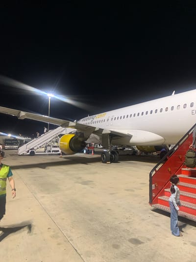 The image depicts an airport tarmac at night, likely at Barcelona–El Prat Airport (BCN) in Sant Boi de Llobregat, Spain, due to the presence of Vueling aircraft, a Spanish low-cost airline headquartered at this airport. A white Vueling airplane is parked on the tarmac, with its wing and engines clearly visible. A mobile airstair is positioned next to the aircraft, with several people ascending and descending it. A young girl in a striped shirt and jeans stands at the bottom of a red mobile boarding stairs, looking up. To the left, a person wearing a bright yellow safety vest stands on the tarmac, appearing to be airport personnel. In the background, a bus and other airport equipment are visible under the artificial lighting of the tarmac. The scene is illuminated by bright floodlights, casting long shadows on the concrete ground.