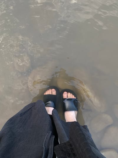 An overhead, medium shot captures a person standing in shallow, murky water with a visible riverbed. The person is wearing black slide sandals and dark, wide-legged pants. The water is partially submerged rocks and debris. The scene appears to be in a natural outdoor setting, possibly a river or stream, with the lighting suggesting daylight. The location is identified as Velykyi Bychkiv, Ukraine.