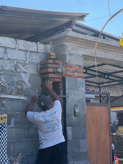A person, wearing a white t-shirt with a blue elephant graphic and text, and a dark baseball cap, is performing masonry work on a wall. The individual is laying bricks, using string to guide the placement. The scene appears to be an outdoor construction site, likely in Sukawati, Indonesia, given the context. The wall is made of concrete blocks, and some sections are already plastered. There are also exposed red bricks in various stages of being laid or dismantled. The background shows elements of a building, including a corrugated metal roof with a lattice structure, and a wooden gate. A light fixture is visible on the gray stone portion of the wall. The lighting suggests it is daytime, with bright sunlight casting shadows. The person's focused posture indicates an active work environment. Visible text on the t-shirt reads "PT USAHA SUKSES MANDIRI" and "MILL & CALCIUM".